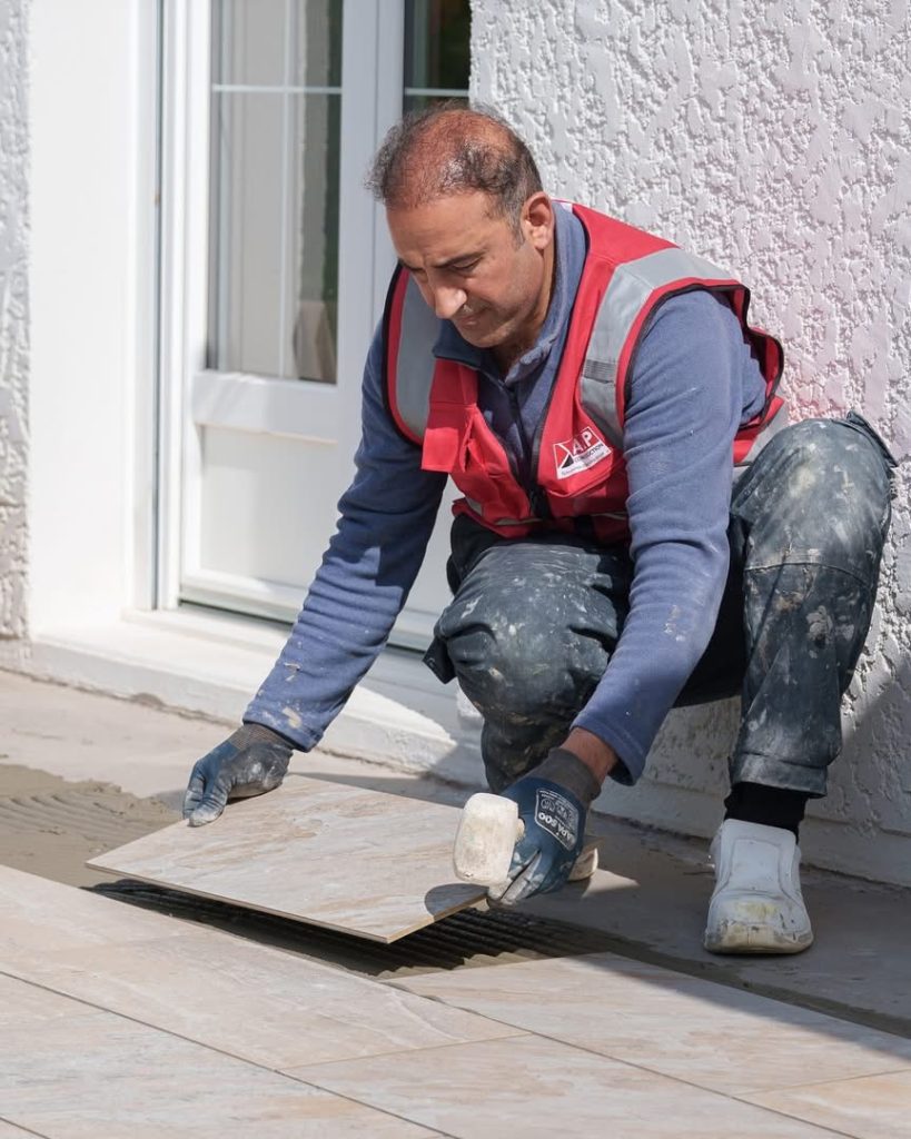 Un carreleur ALP positionne un carreau au sol devant une porte-fen&ecirc;tre, sur une terrasse en cours de r&eacute;alisation.