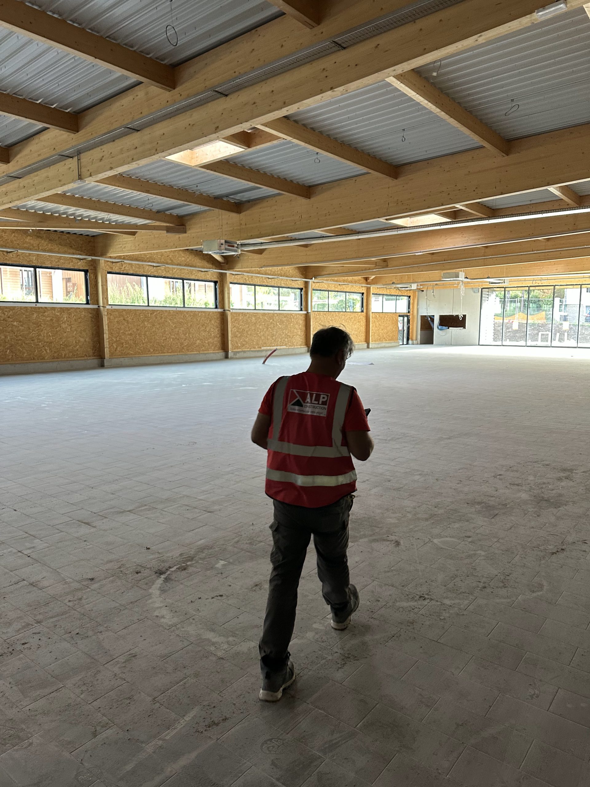 Un salari&eacute; ALP vu de dos observe l&rsquo;avancement des travaux dans un grand b&acirc;timent &agrave; charpente apparente.