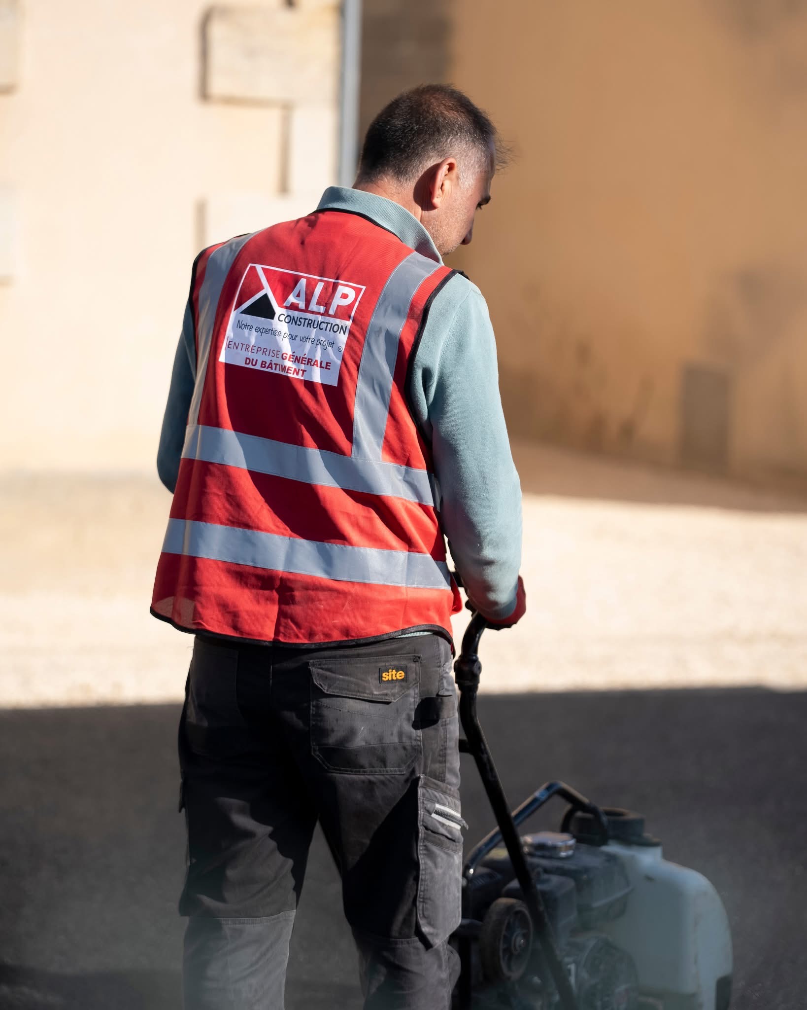 Un salarié ALP vu de dos conduit un outil de compactage sur un sol de chantier.