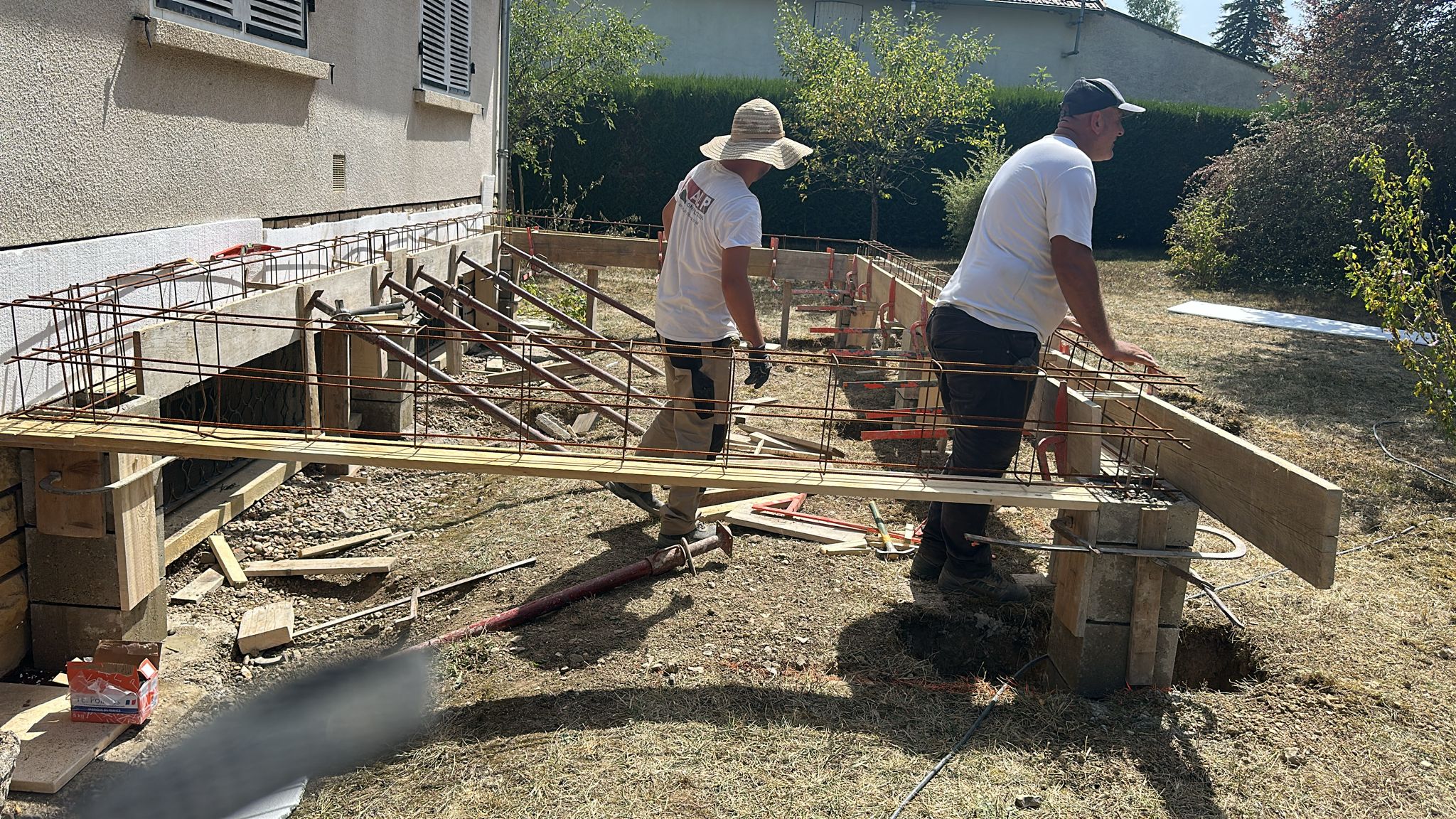 Deux ouvriers ALP transportent une armature m&eacute;tallique au-dessus des fondations d&rsquo;un chantier.