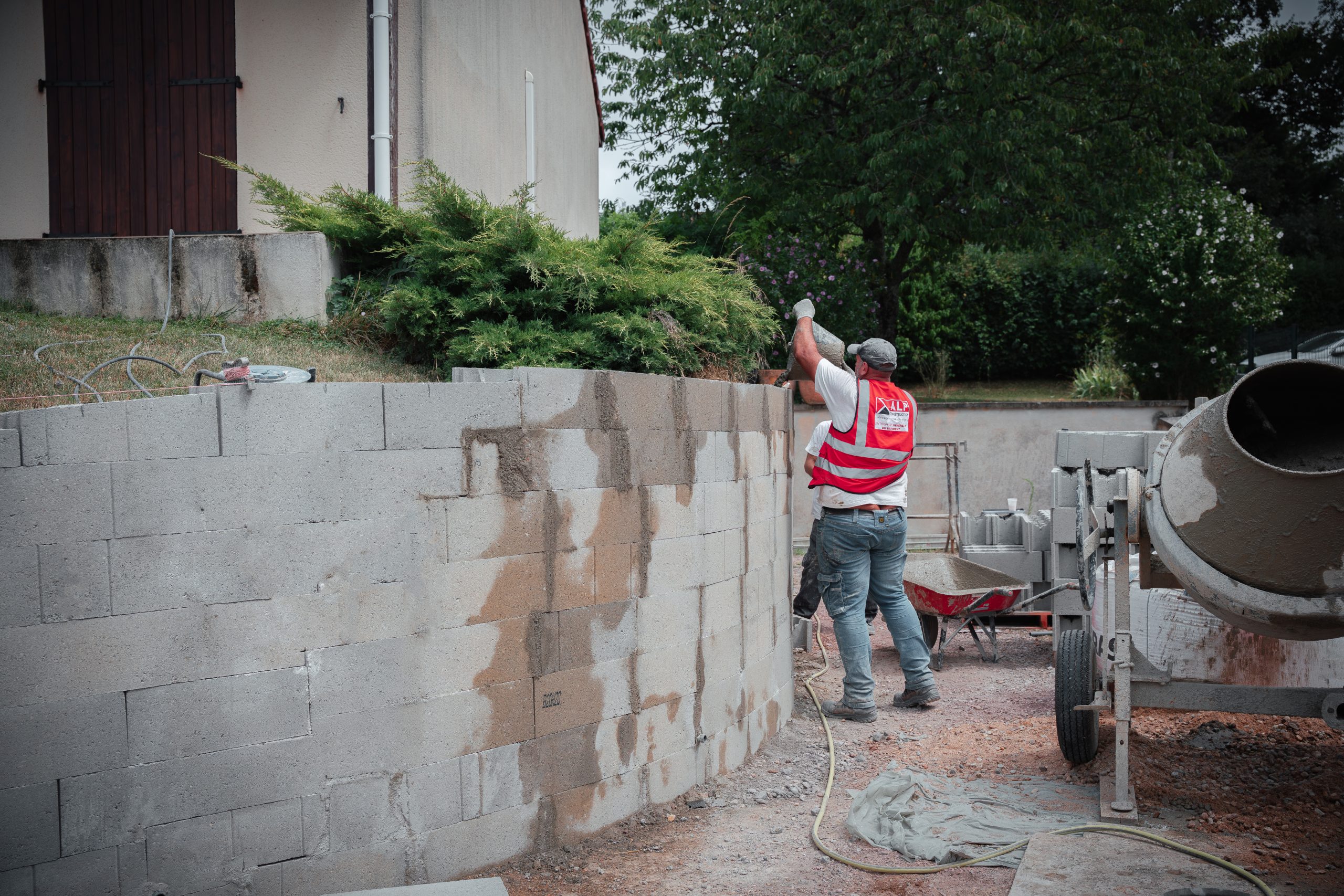 Un maçon ALP monte un mur en parpaings, avec une bétonnière et des matériaux stockés à proximité.