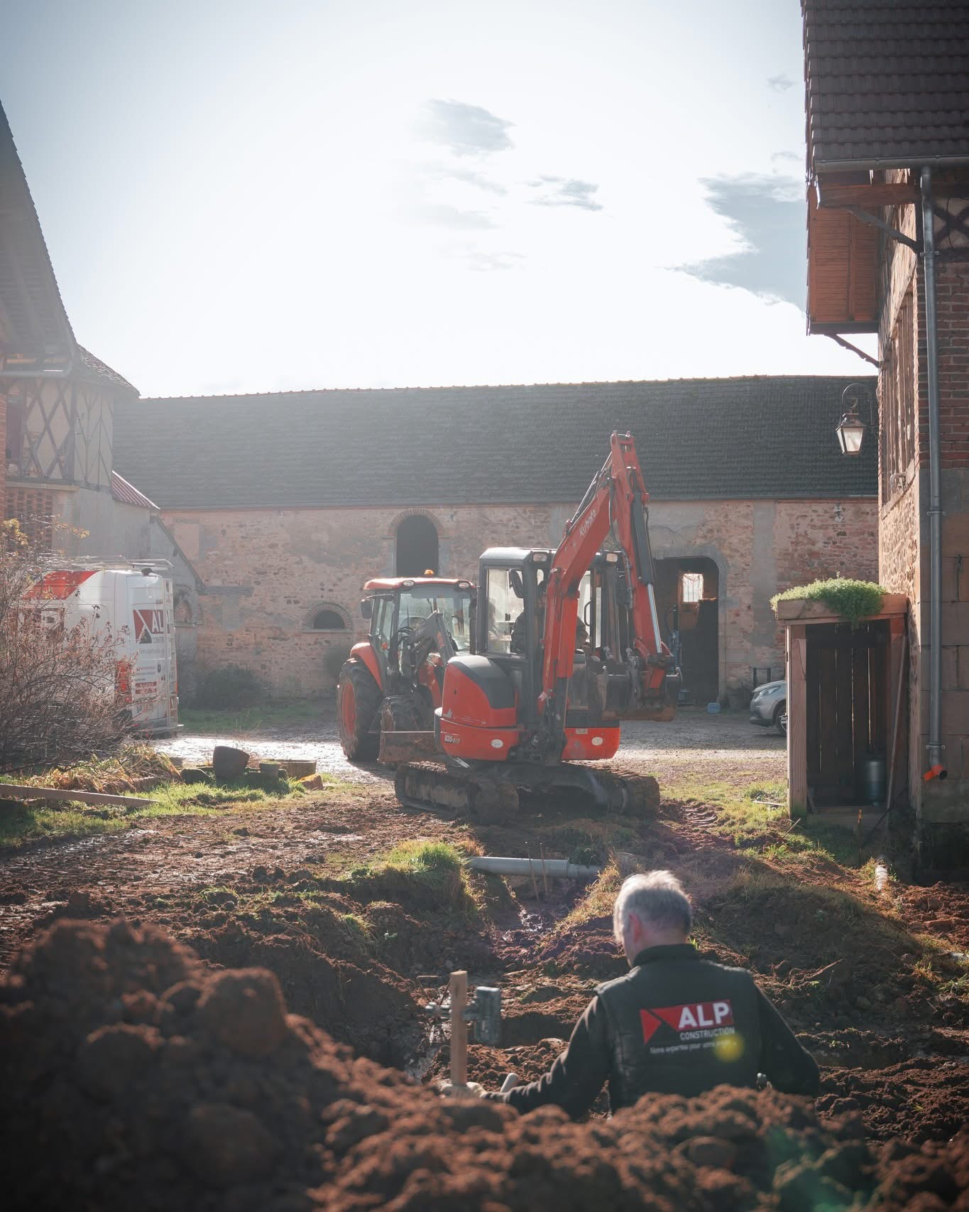 Une mini-pelle réalise un terrassement devant une maison, accompagnée par des salariés sur le chantier.