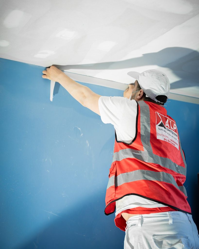 Un peintre ALP vu de dos r&eacute;alise une finition &agrave; la jonction d&rsquo;un mur bleu et d&rsquo;un plafond blanc.